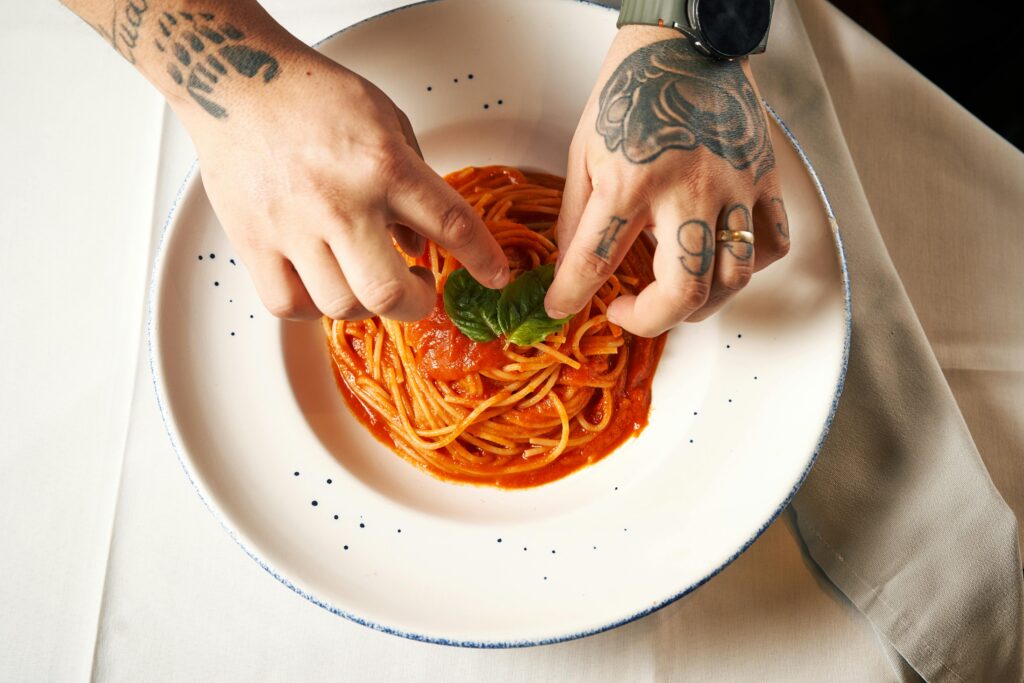 Tattooed hands adding basil to spaghetti with tomato sauce, offering a culinary touch.