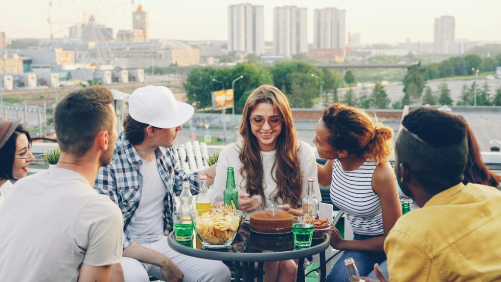 A cheerful group of friends enjoys a celebration with cake on a sunny rooftop.
